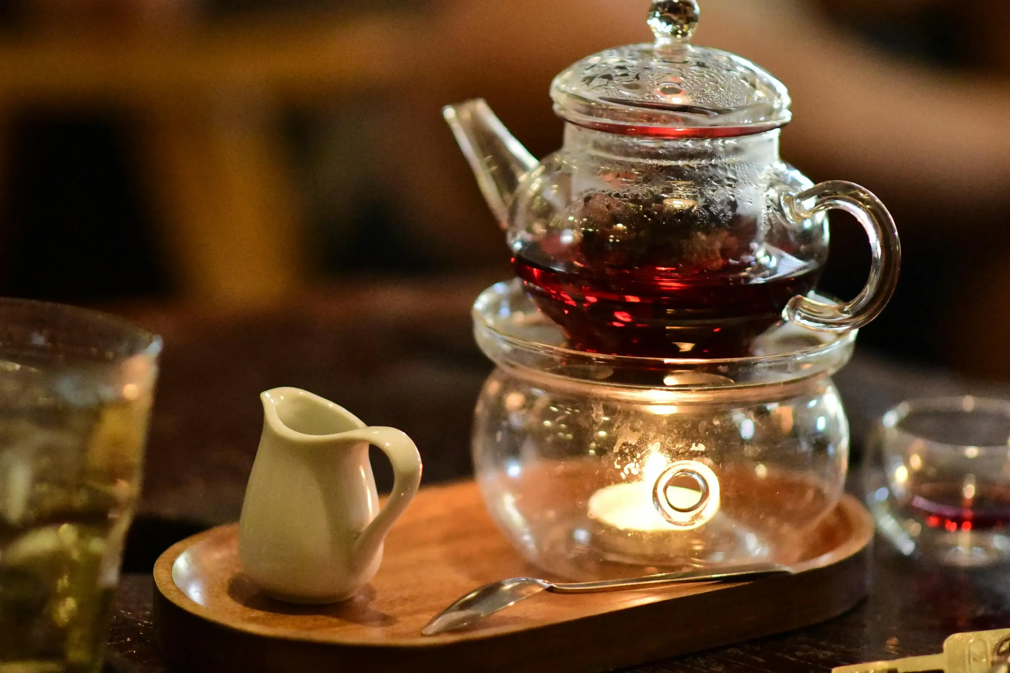 A clear glass teapot sits atop a glass warmer, illuminated by the soft glow of a small candle flickering beneath the dark red tea. The set is presented on a wooden tray alongside a petite white ceramic creamer and a delicate silver spoon.
