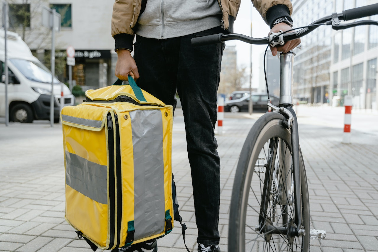 Entregador de bike segurando bicicleta e caixa térmica. Foto: Pexels