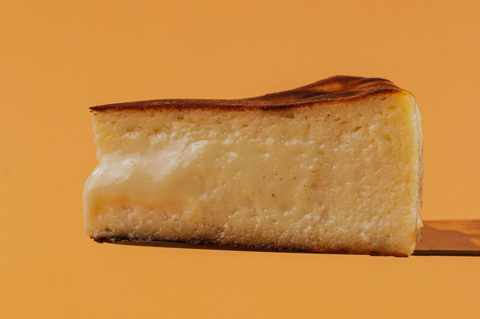 A slice of creamy, golden-brown cheesecake with a caramelized top rests on a spatula against a warm orange background, evoking a tempting and cozy feel.