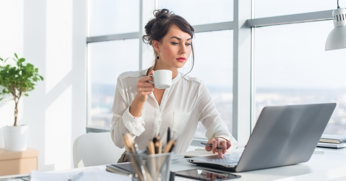 A focused businesswoman sipping coffee while researching tax saving strategies on her laptop in a bright, spacious office.