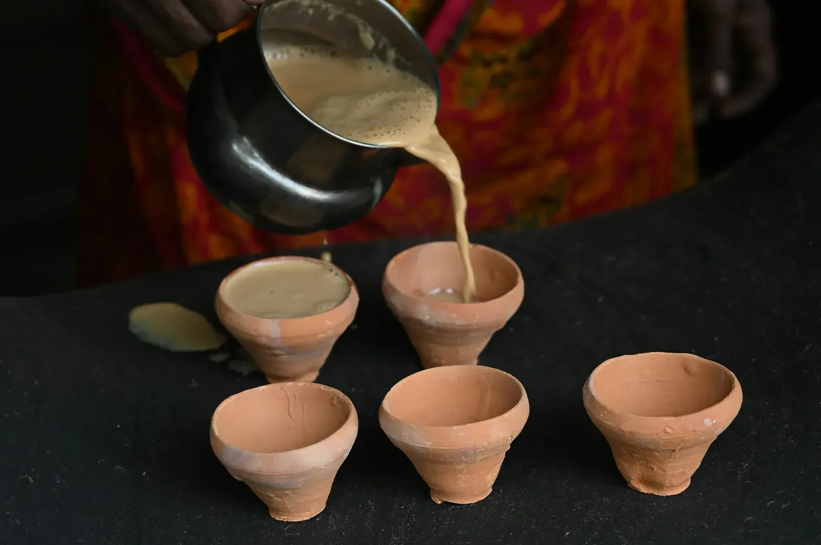 A person, seen from the torso down, pours hot, frothy masala chai from a metallic pot into small, traditional unglazed clay cups arranged on a dark, flat surface, emphasizing the authentic method of serving street-style tea.