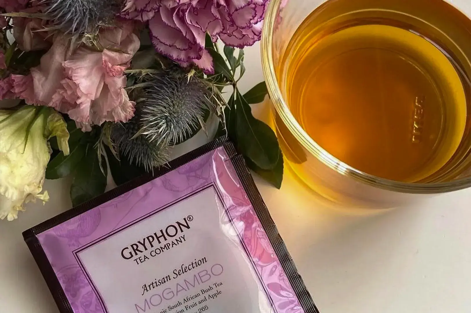 A high-angle, close-up composition showcasing a dark purple and pink sachet of Gryphon Tea Company's Artisan Selection "Mogambo" tea, which features an organic South African bush tea blend with passion fruit and apple. The individual tea packet rests diagonally on a white surface next to a glass cup filled with clear, amber-colored brewed tea seen from above. A delicate bouquet of textured, dusty-purple carnations and spiky, bluish sea holly thistles decorates the top-left corner, adding a soft, elegant floral element to the scene.