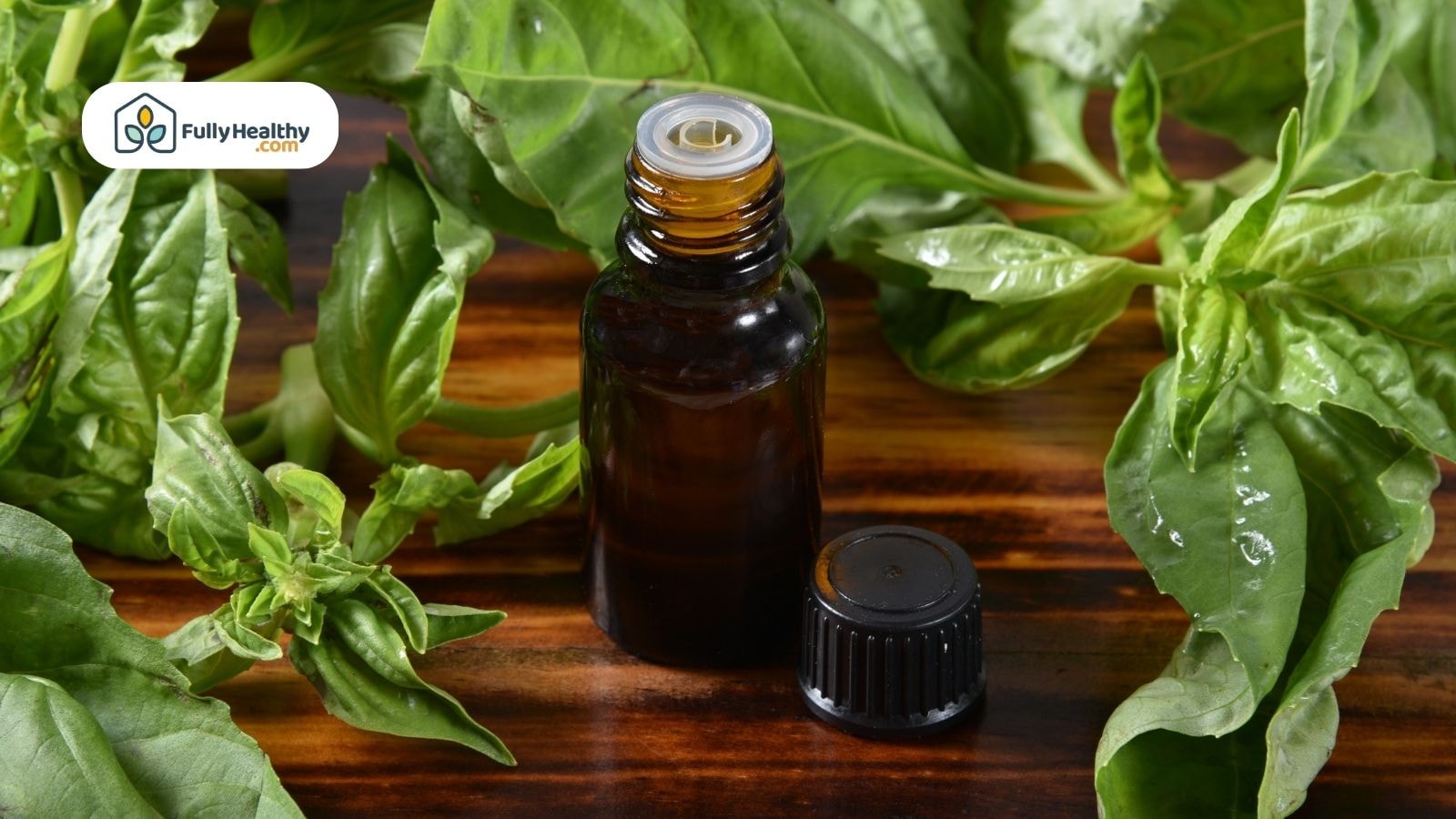 Brown glass bottle of basil essential oil surrounded by fresh basil leaves on a wooden surface