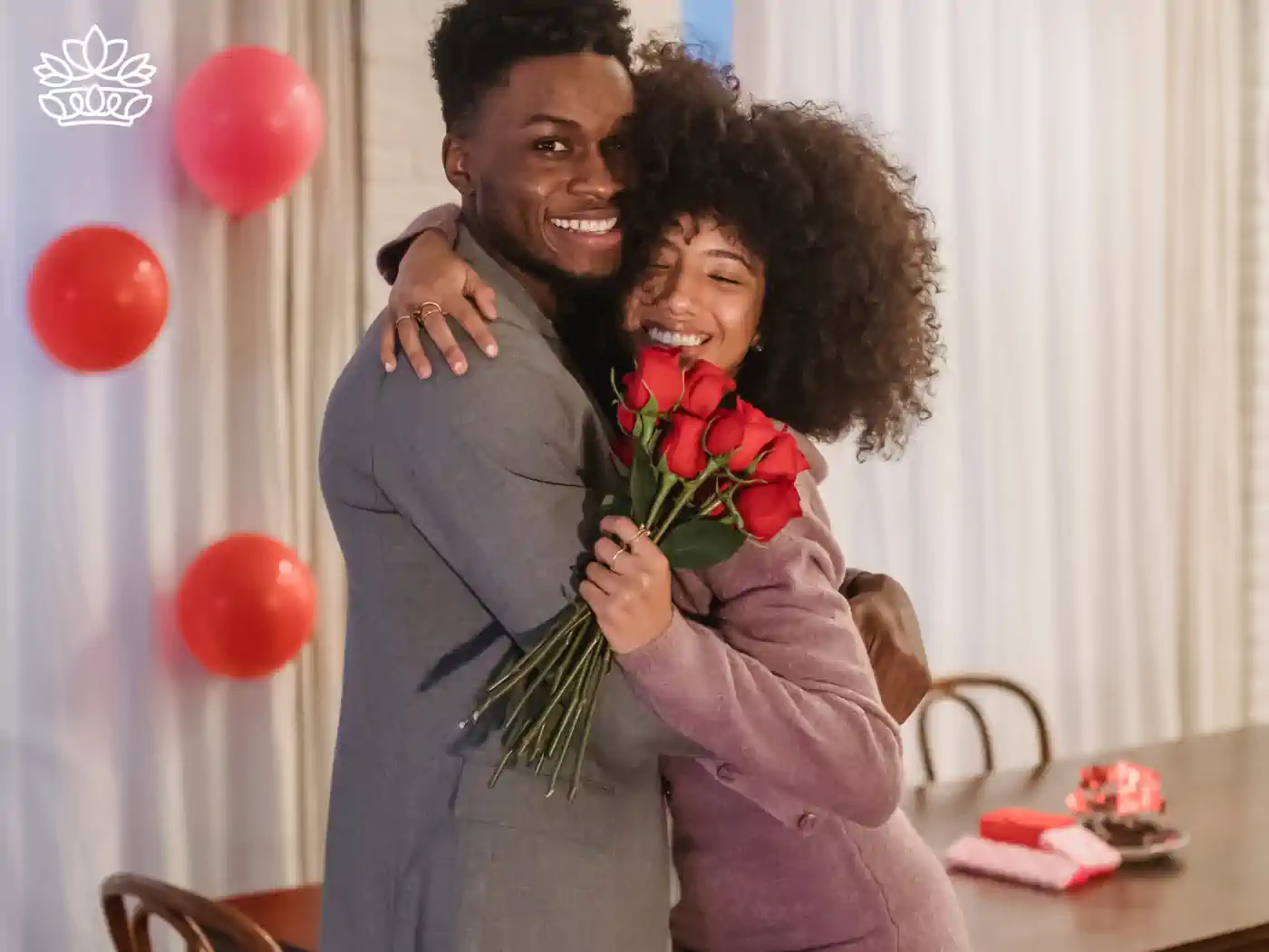 A joyful embrace between a couple as the woman receives a bouquet of red roses, with red balloons and Valentine's decor in the background. Fabulous Flowers and Gifts delivered with heart. Valentine's Day Flowers.