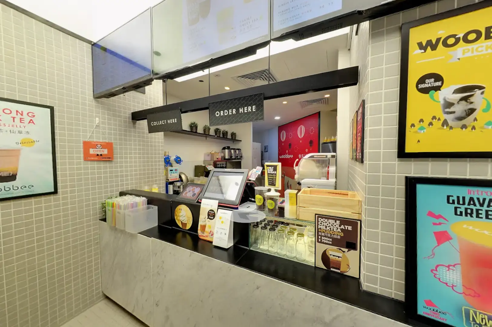 A modern Woobbee bubble tea store interior with light tiled walls, black service counters labeled "ORDER HERE" and "COLLECT HERE," digital menu boards overhead, and promotional posters showcasing signature drinks like Guava Green Tea and Double Chocolate Milk Tea.