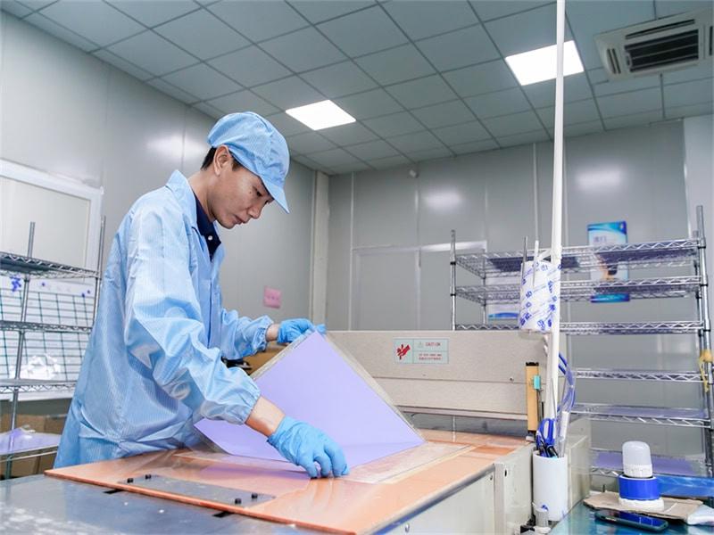 A factory worker in a blue cleanroom uniform, cap, and gloves carefully handling a large sheet of purple thermal material on a production line. The setting is a bright, organized industrial facility with stainless steel shelving and specialized machinery.