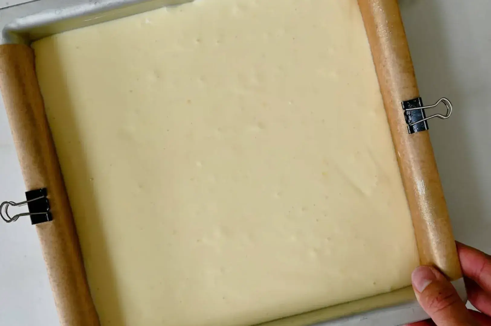 A baking pan filled with a smooth, creamy cheesecake batter, secured with clips and wooden rolling pins on either side.