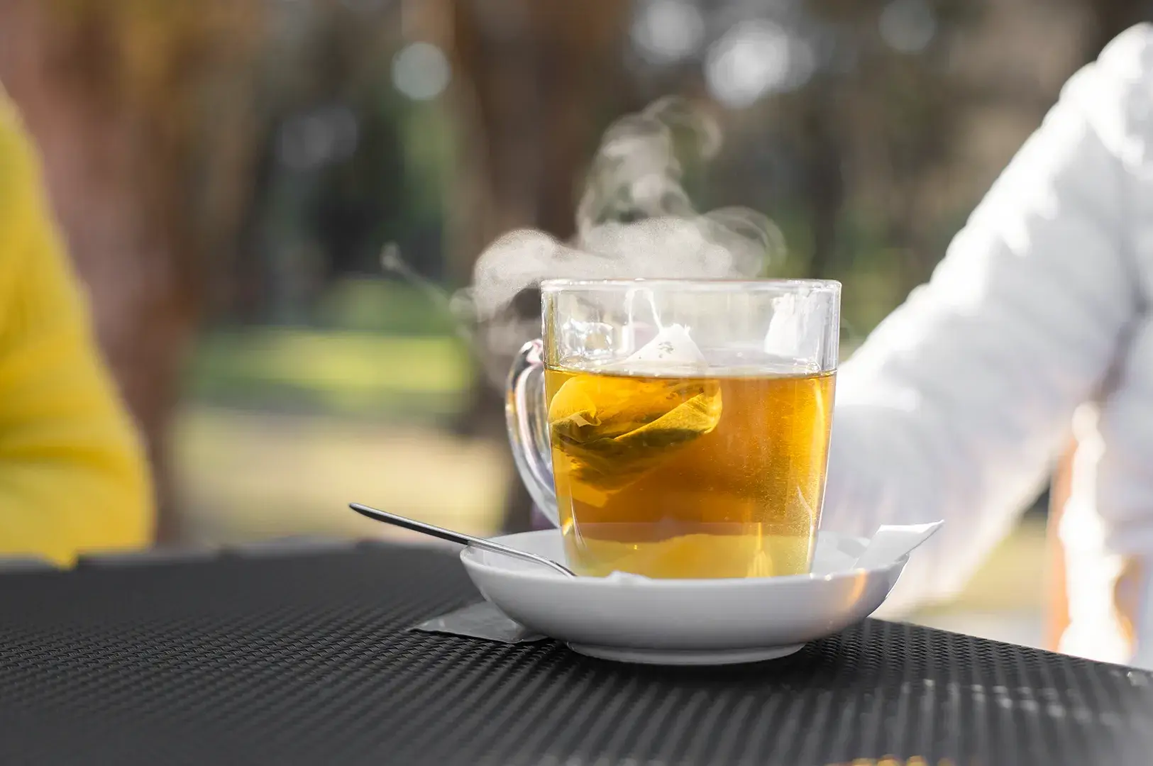 Steaming glass mug of tea with a tea bag on an outdoor table.