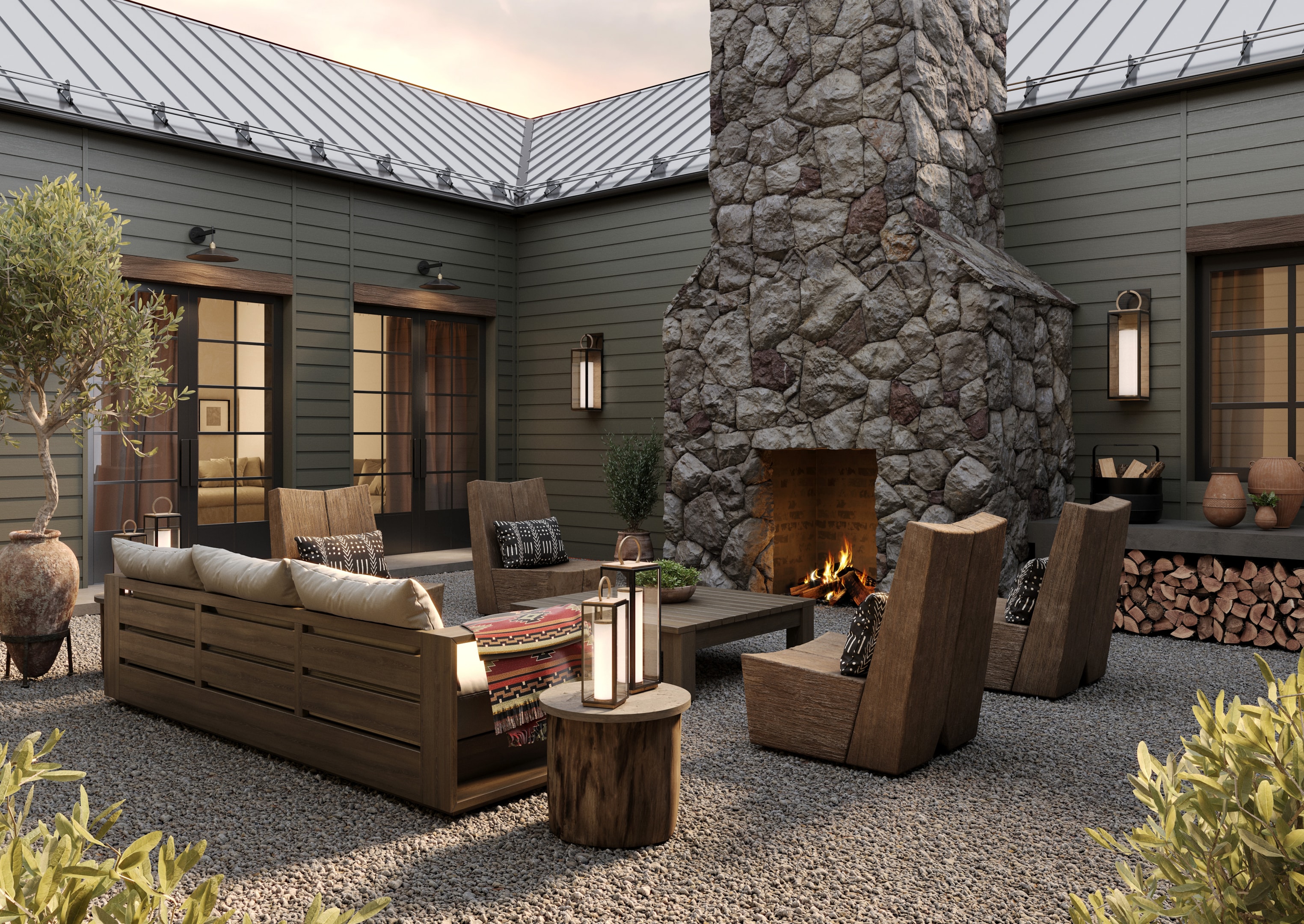 Outdoor patio with a stone fireplace, wooden seating, lantern lighting, and a rustic coffee table on gravel, surrounded by greenery and farmhouse-style architecture.