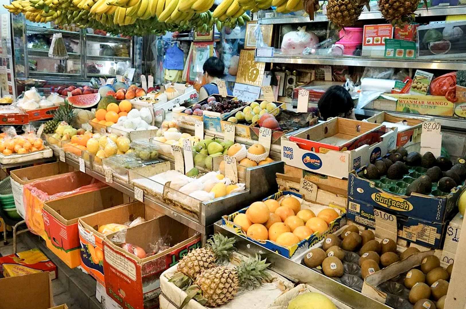A vibrant market stall filled with a wide variety of fresh fruits, including bananas, pineapples, citrus, and other produce arranged in boxes and trays.