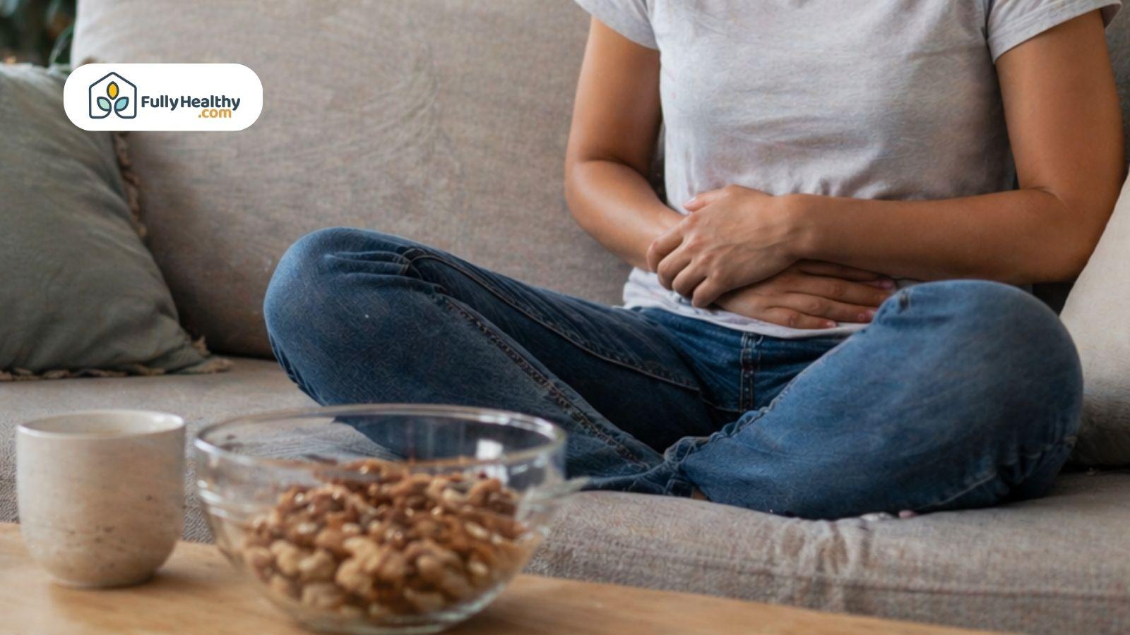 Person sitting on couch holding stomach with bowl of mixed nuts on table.