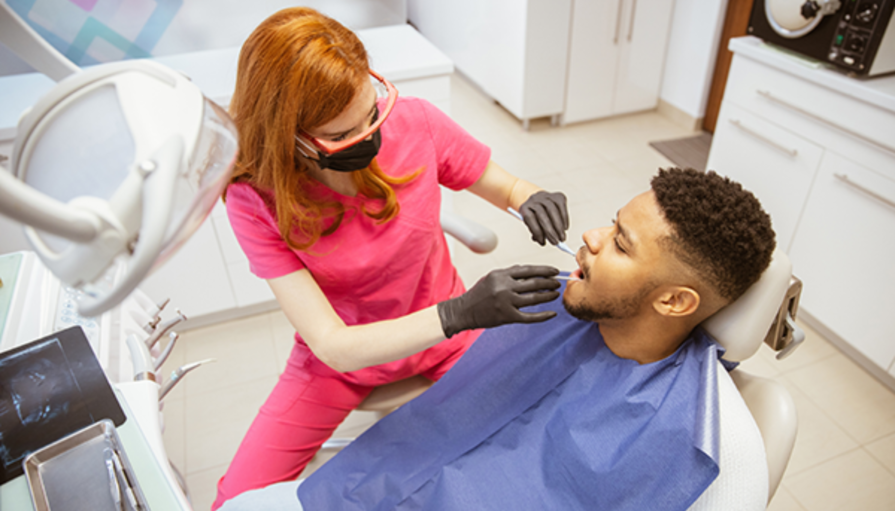 Tannspesialist på Eiksmarka – Når trenger du én? 15 Tannpleier i rosa uniform undersøker en manns tenner på tannlegekontor, pasienten sitter i behandlingsstol med blå beskyttelsesduk.