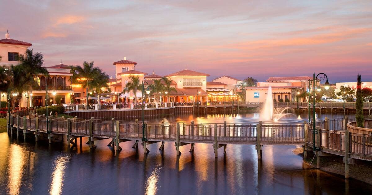 The image showcases a picturesque view of Coconut Point's lakeside boardwalks, lined with palm trees and inviting outdoor dining areas, perfect for enjoying meals like thin crust pizzas and salads at local restaurants such as Divieto Ristorante. This scenic spot in Estero, Florida, is an ideal hub for families and residents to relax and savor the taste of delicious food while taking in the beautiful surroundings.