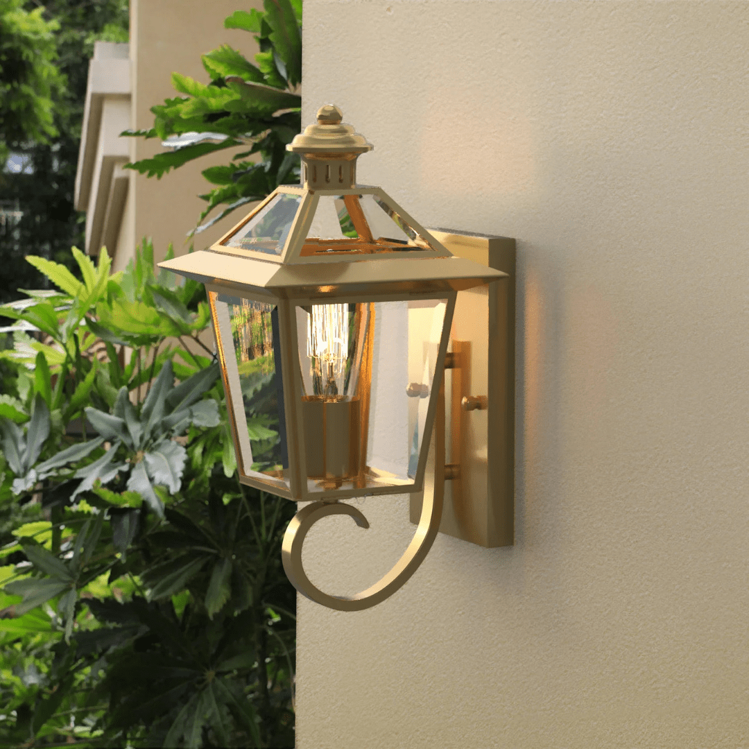 Gold outdoor wall lantern with clear glass panels and a glowing filament bulb, mounted on a light-colored wall near green foliage.