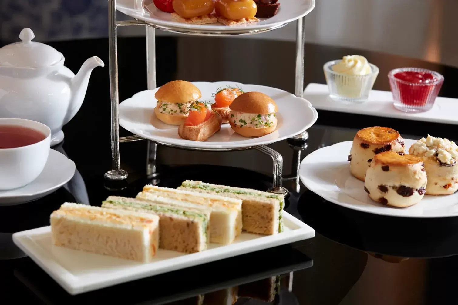 A three-tier silver stand holds an elegant afternoon tea service featuring assorted finger sandwiches, savory brioche sliders, and delicate pastries. Beside it, a white teapot and cup are paired with fresh fruit scones, clotted cream, and strawberry jam on a sleek black tabletop.
