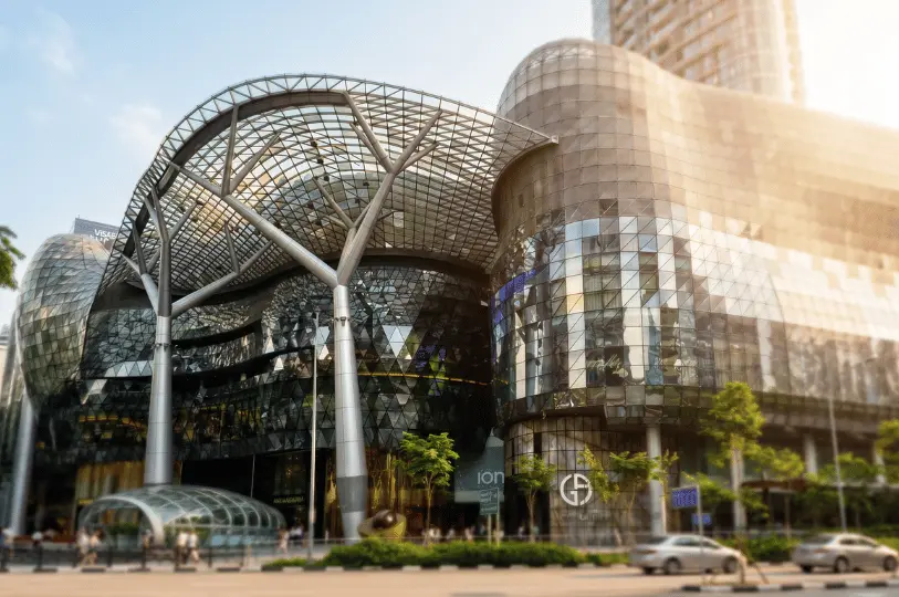 The modern exterior of the ION Orchard shopping mall in Singapore, highlighting its iconic curved glass facade and striking tree-like steel canopy structure against a bright sky.