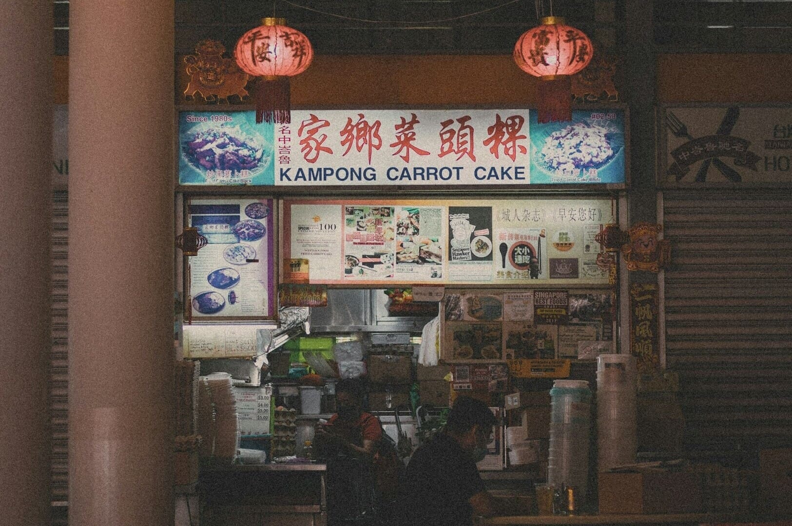  A man sitting at a table outside Singapore Hawkers with a vibrant street scene in the background.