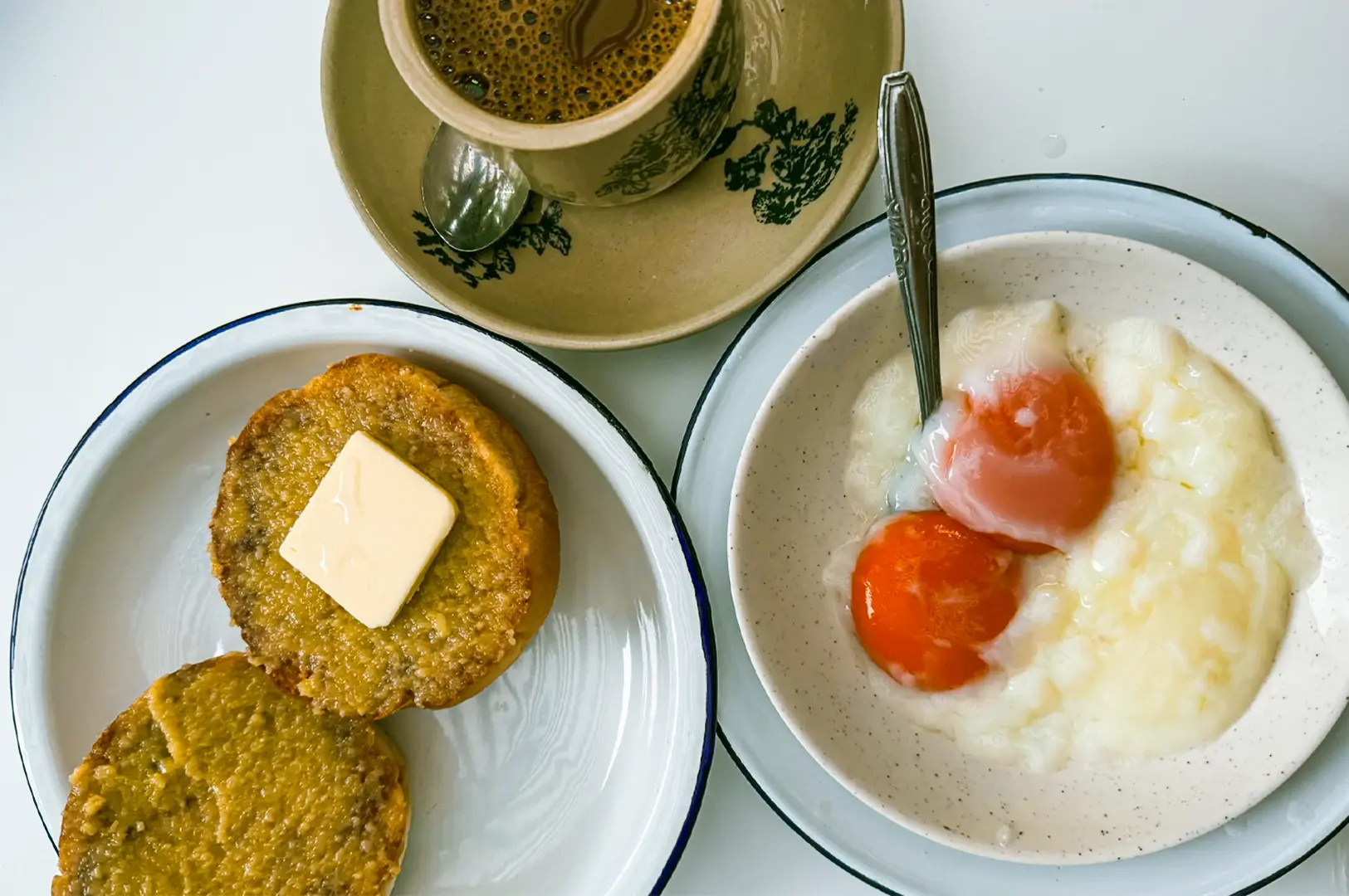 Overhead top‑down shot of a traditional Singapore breakfast featuring soft‑boiled eggs in a bowl, buttered kaya toast biscuits on a plate, and a cup of local coffee served on a light tabletop.