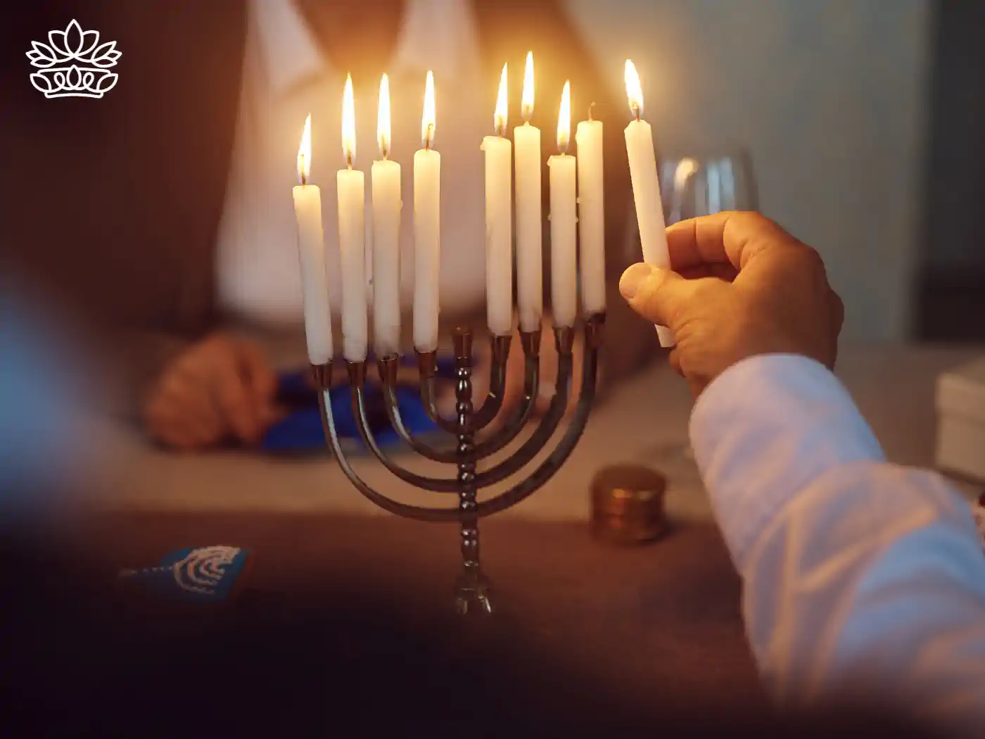 A hand lighting a white candle on a menorah, with eight other candles already lit, in a warm and cosy setting. Fabulous Flowers and Gifts - Hanukkah Flowers.