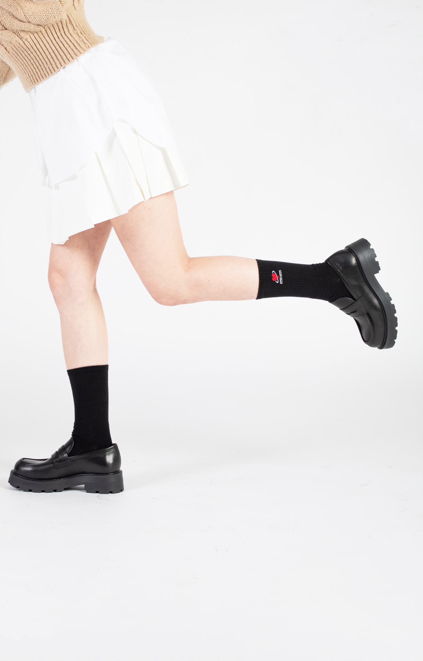 A female model wears black leather loafers with a pair of black crew socks with a heart embroidered in red thread.