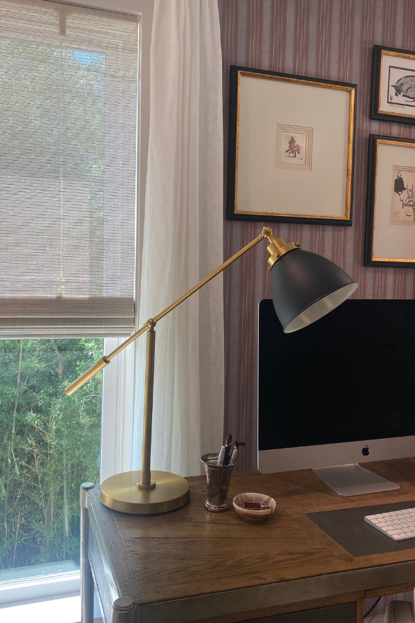 A wooden desk with a brass and black desk lamp, computer monitor, and framed artwork on a wallpapered wall, styled as a cozy home office corner.