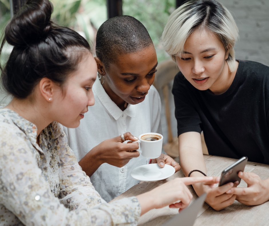 three women sit at a table drinking coffee and looking at one person's phone