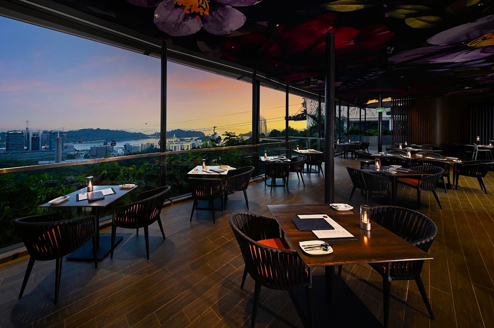 Modern restaurant with floral ceiling, wooden tables, and candlelight. Overlooks cityscape at sunset, creating a warm and inviting ambiance.