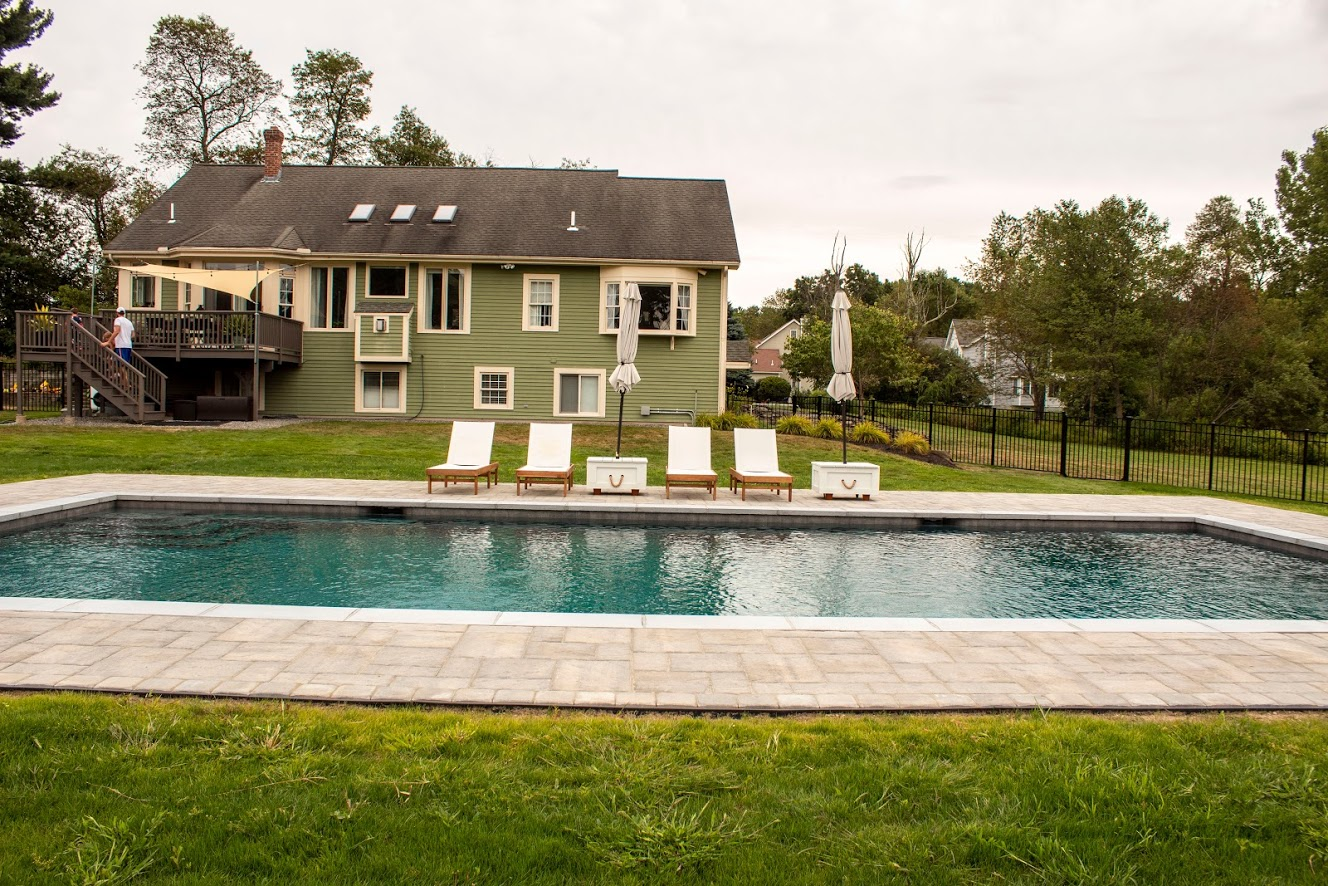 A backyard with a rectangular swimming pool, four lounge chairs, and a green house with a deck and stairs in the background.