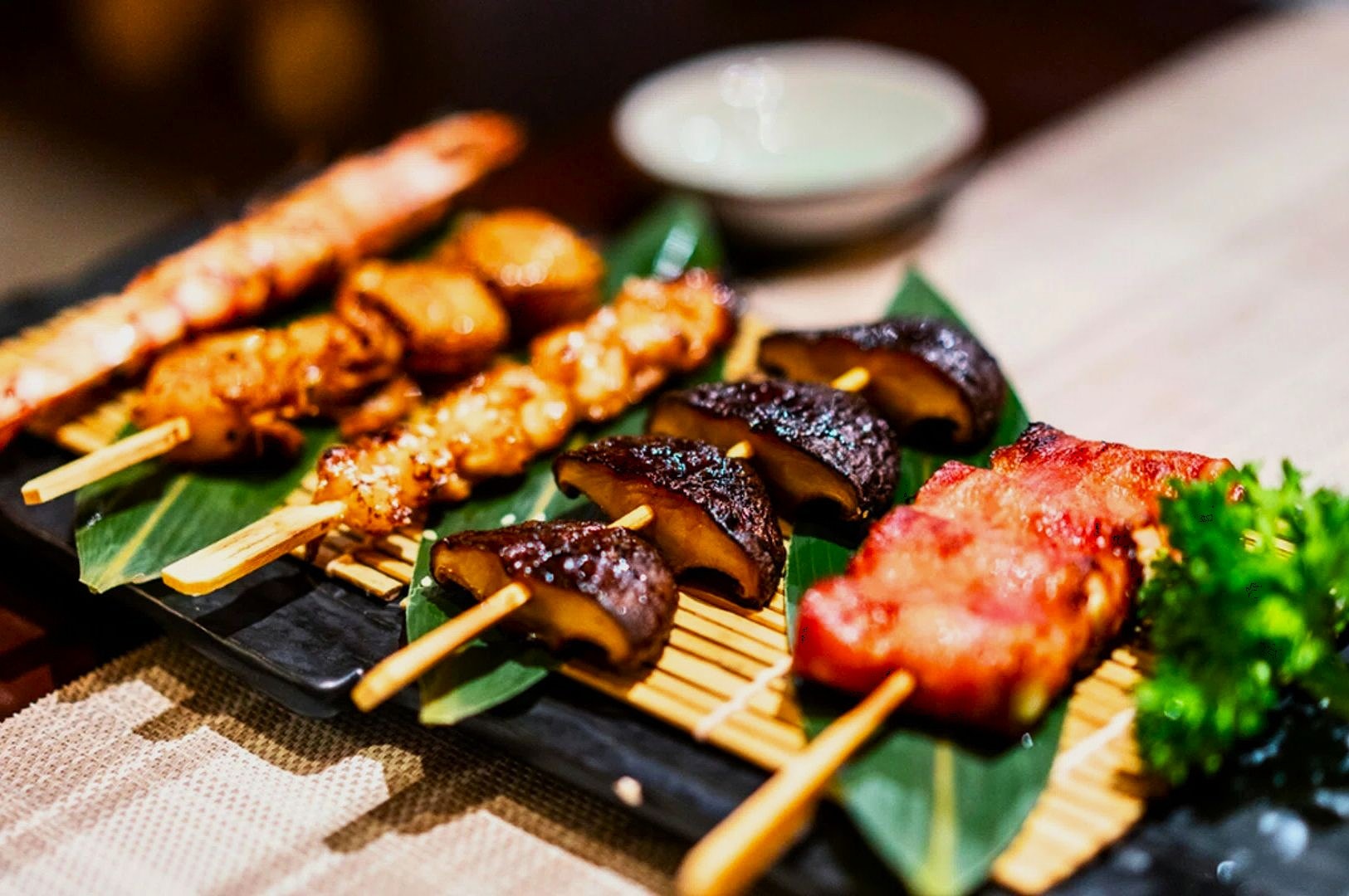 Assorted skewers with shrimp, chicken, mushrooms, and bacon-wrapped items on banana leaves, garnished with greens. Elegant presentation on bamboo.