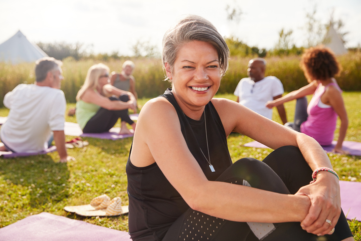 An image of a smiling mature woman sitting on a yoga mat in the park.