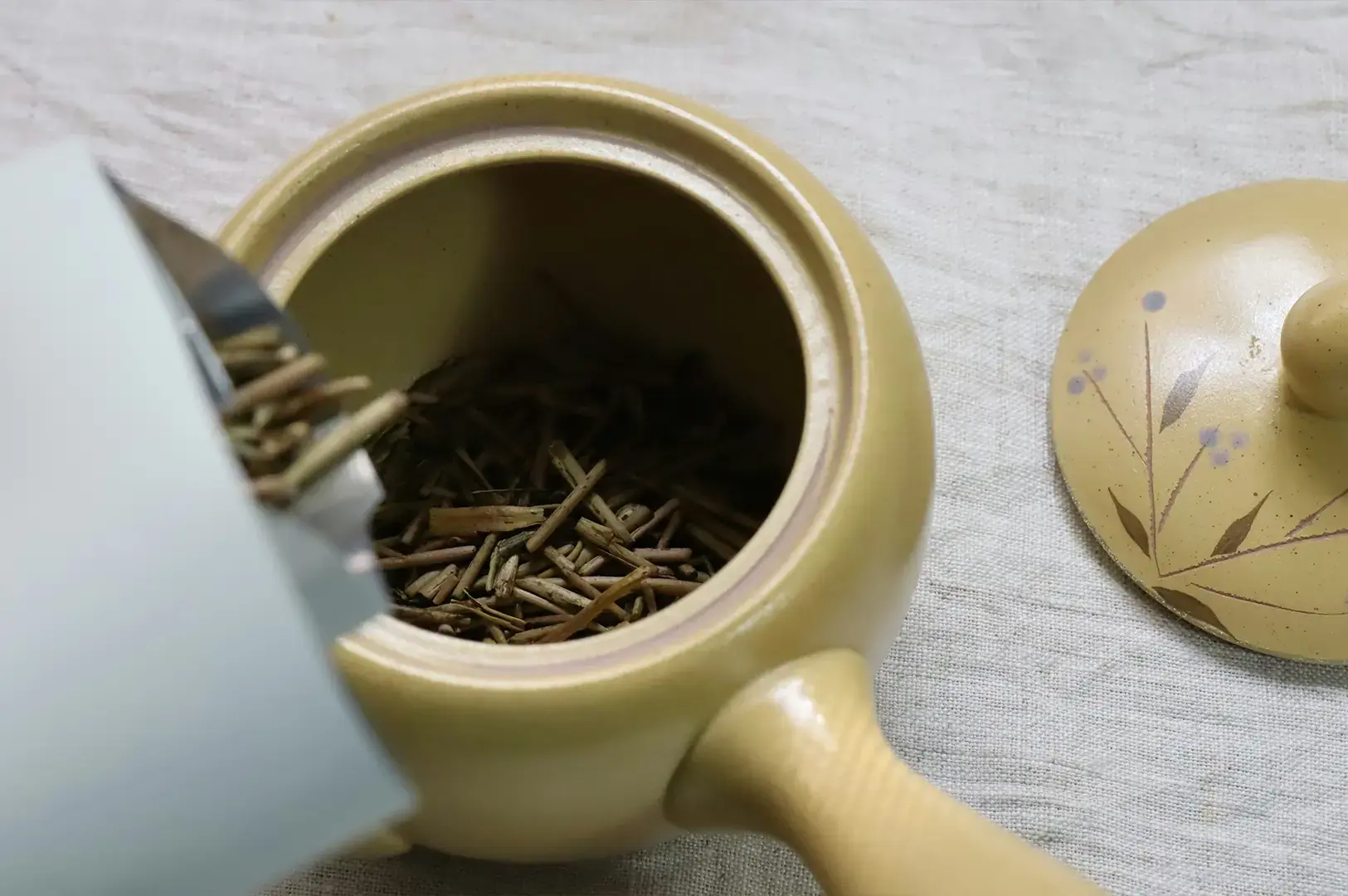 Dry, needle-like tea leaves are being poured from a light-colored paper bag into a yellow ceramic teapot, with the lid of the teapot resting nearby on a textured beige fabric surface.