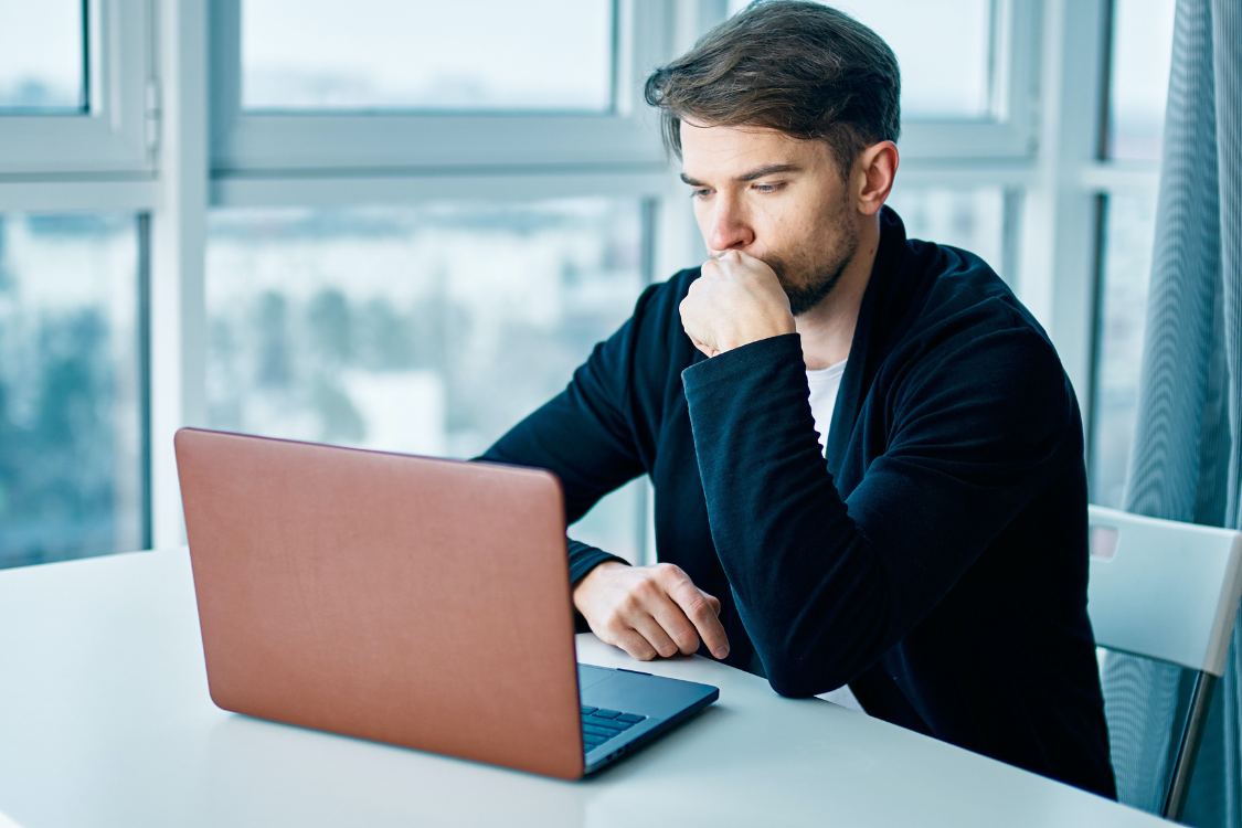Man thinking while working on a laptop, weighing options between selling his business alone or hiring a broker.