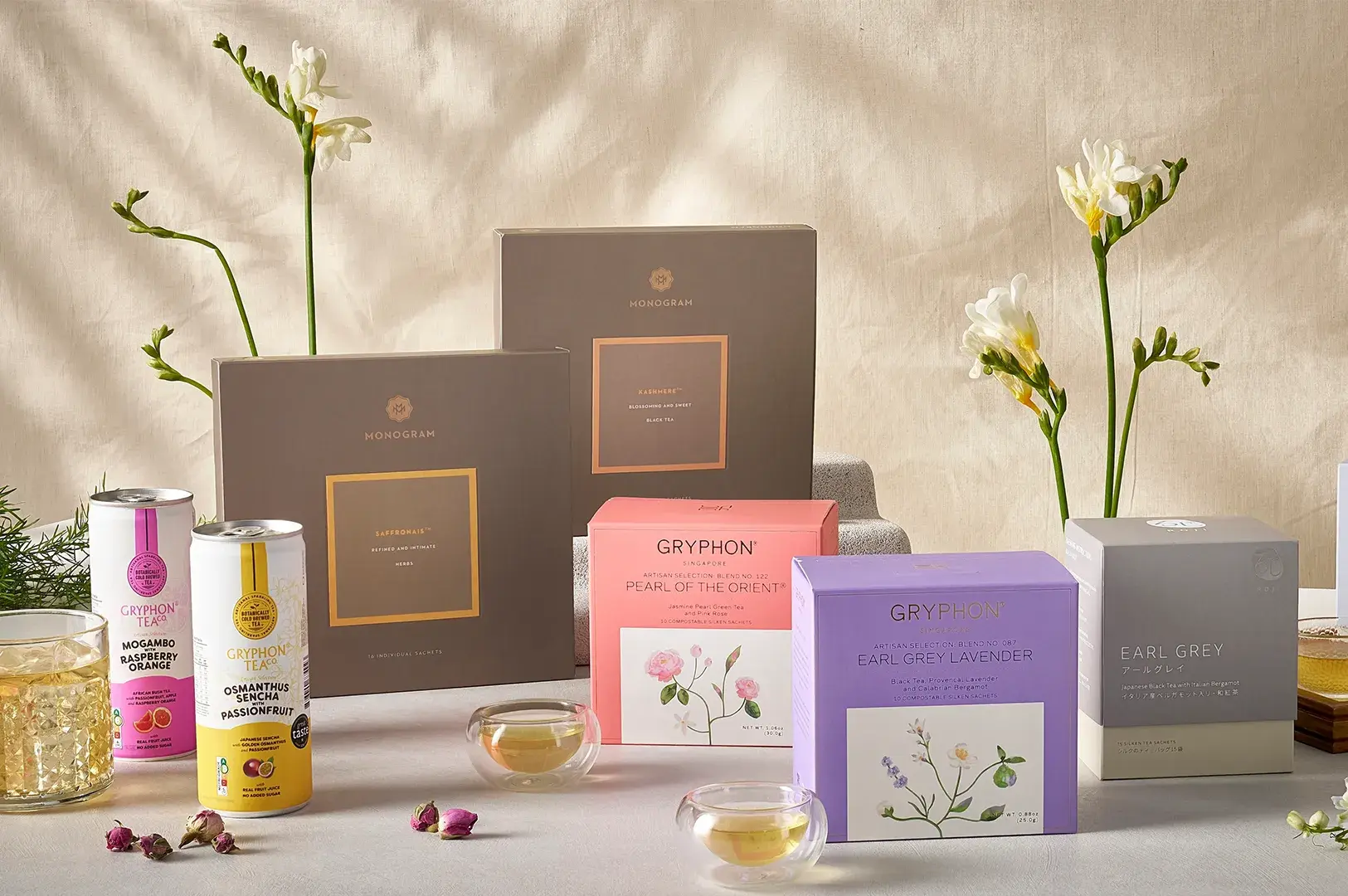 Product display featuring Gryphon Tea Co. and Monogram tea boxes, canned sparkling teas, small glass tea cups, and white freesias under warm sunlight.