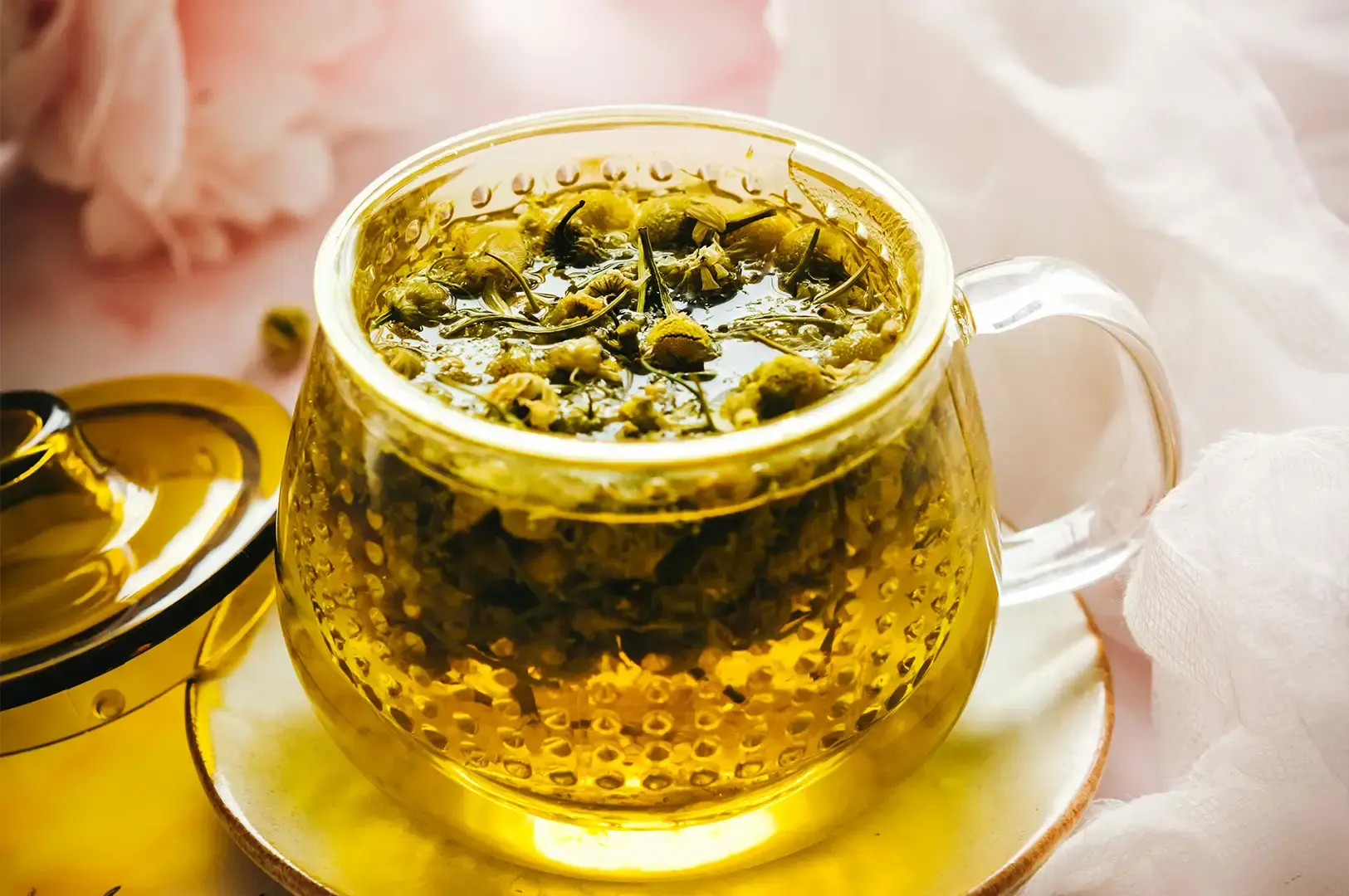 A vibrant, top-down close-up of a clear glass mug filled to the brim with yellow herbal tea and packed tightly with numerous small, steeped chamomile or chrysanthemum flower buds. The mug sits on a matching yellow saucer, positioned against a soft, dreamy background of pink floral elements and draped white translucent cloth, highlighted by bright, warm lighting.