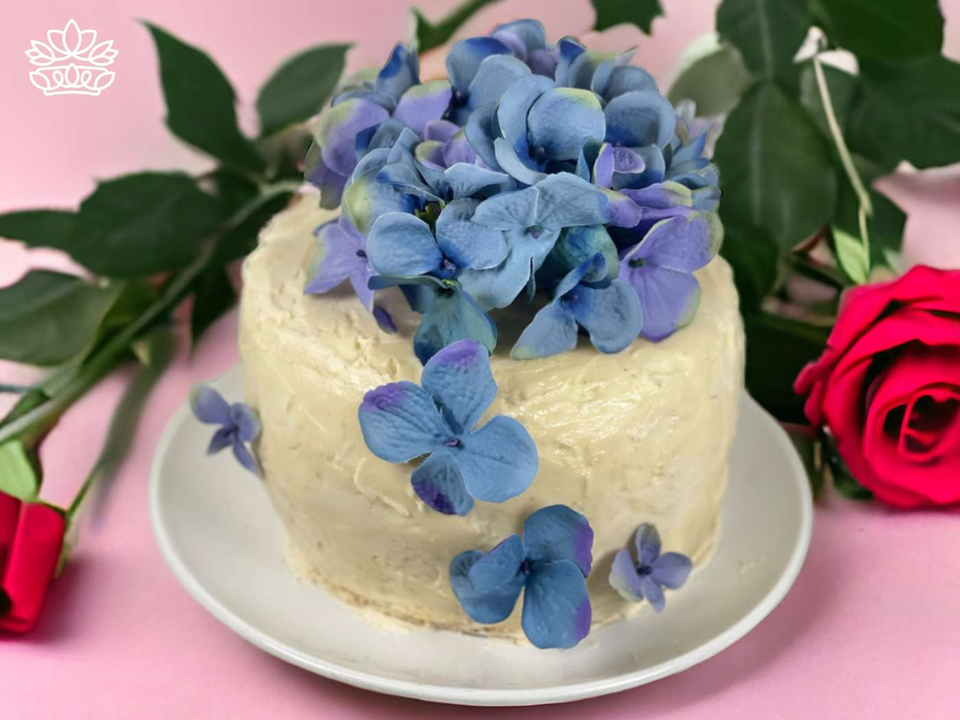 A beautifully frosted cake topped with a cluster of delicate blue hydrangeas, set against a pink background with a vivid red rose in the foreground, reflecting the artistry available at Fabulous Flowers and Gifts.