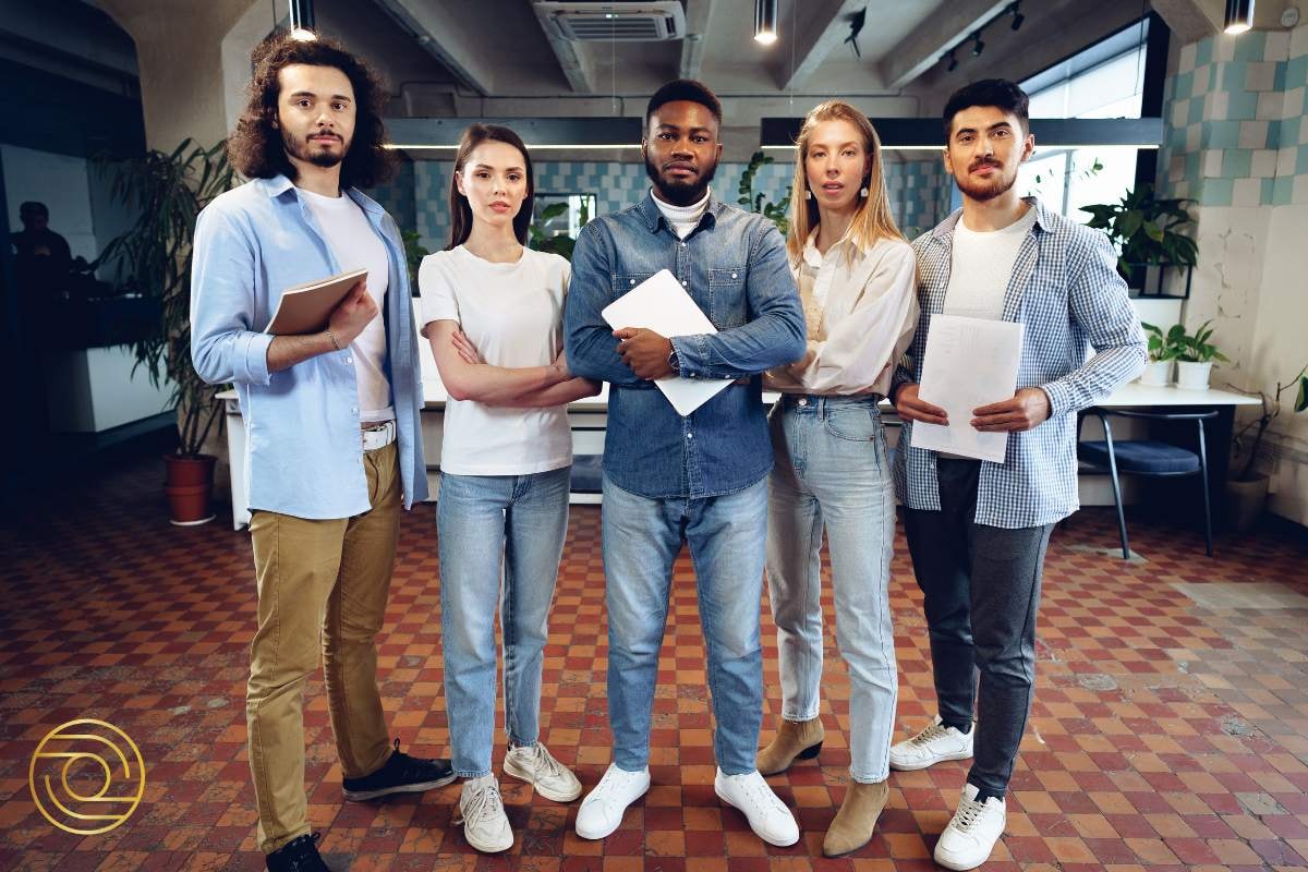 Group of five professionals standing together in a modern office setting, holding documents and looking focused, symbolising readiness and shared vision.