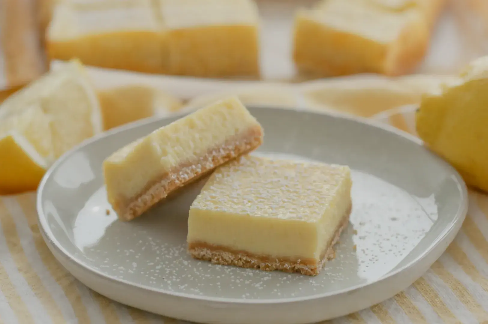 A plate with two lemon bars, one slightly tilted, dusted with powdered sugar, with lemon slices in the background.