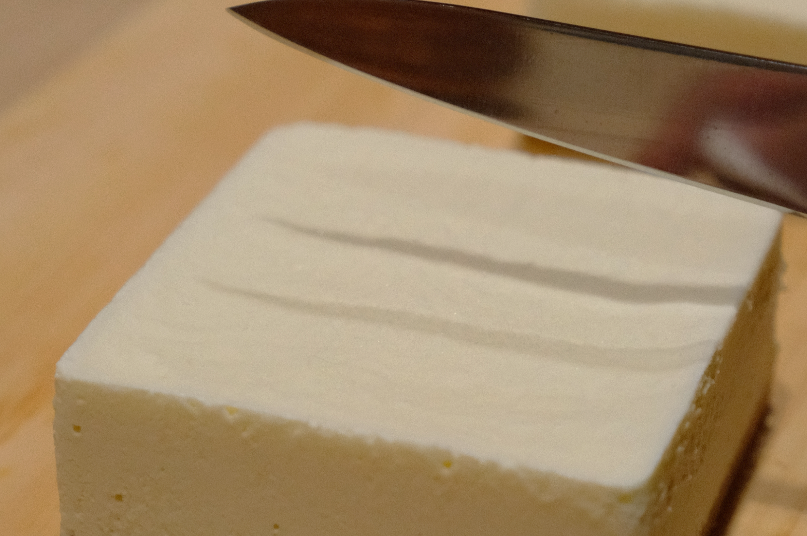 A sharp knife poised above a block of tofu on a wooden cutting board, ready to slice. The scene evokes preparation and attention to detail.