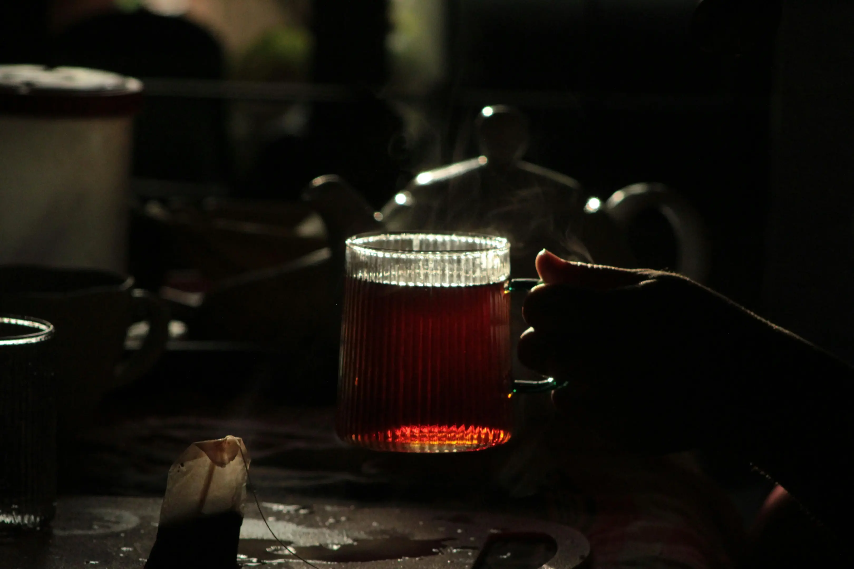 A person holds a ribbed, transparent glass mug filled with a dark amber tea, which catches the light in a dimly lit setting. In the soft, shadowy background, a teapot and an used tea bag are subtly visible on the table.