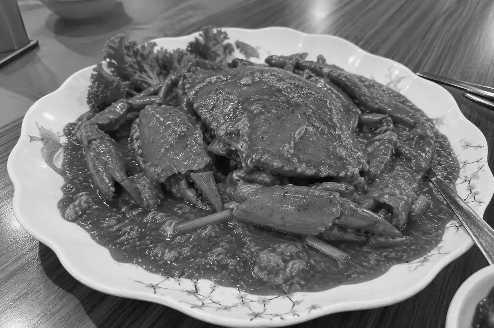 A whole chili crab covered in thick savory sauce, served on a white decorative plate with floral patterns on a wooden restaurant table.