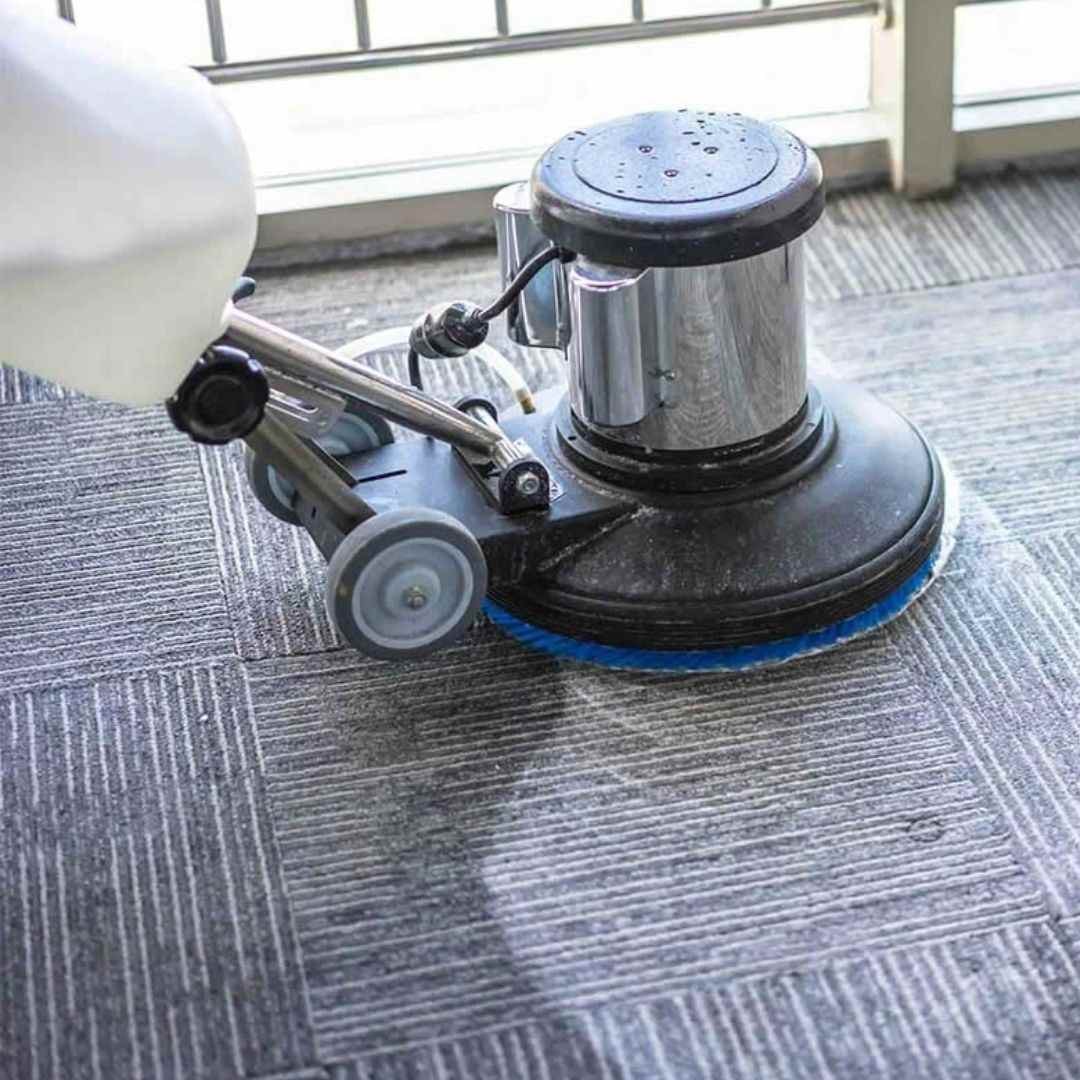 Close-up of a heavy-duty floor buffer machine scrubbing a grey patterned carpet near a window, highlighting the deep-cleaning process and texture of the carpet fibers.