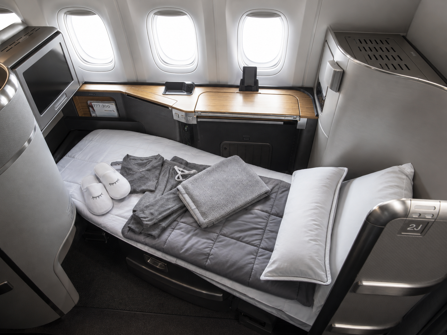 Luxury business class suite with a fully flat bed, white pillows, grey bedding, sleepwear and slippers beside large aircraft windows.