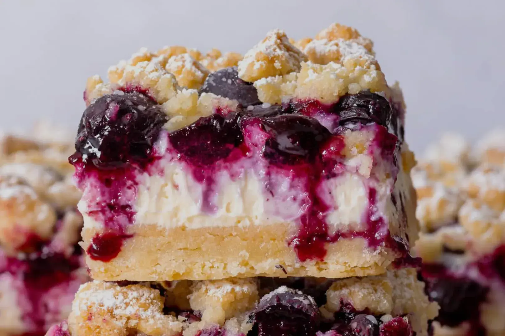 A close-up of layered dessert bars with a creamy filling and a berry topping, dusted with powdered sugar.