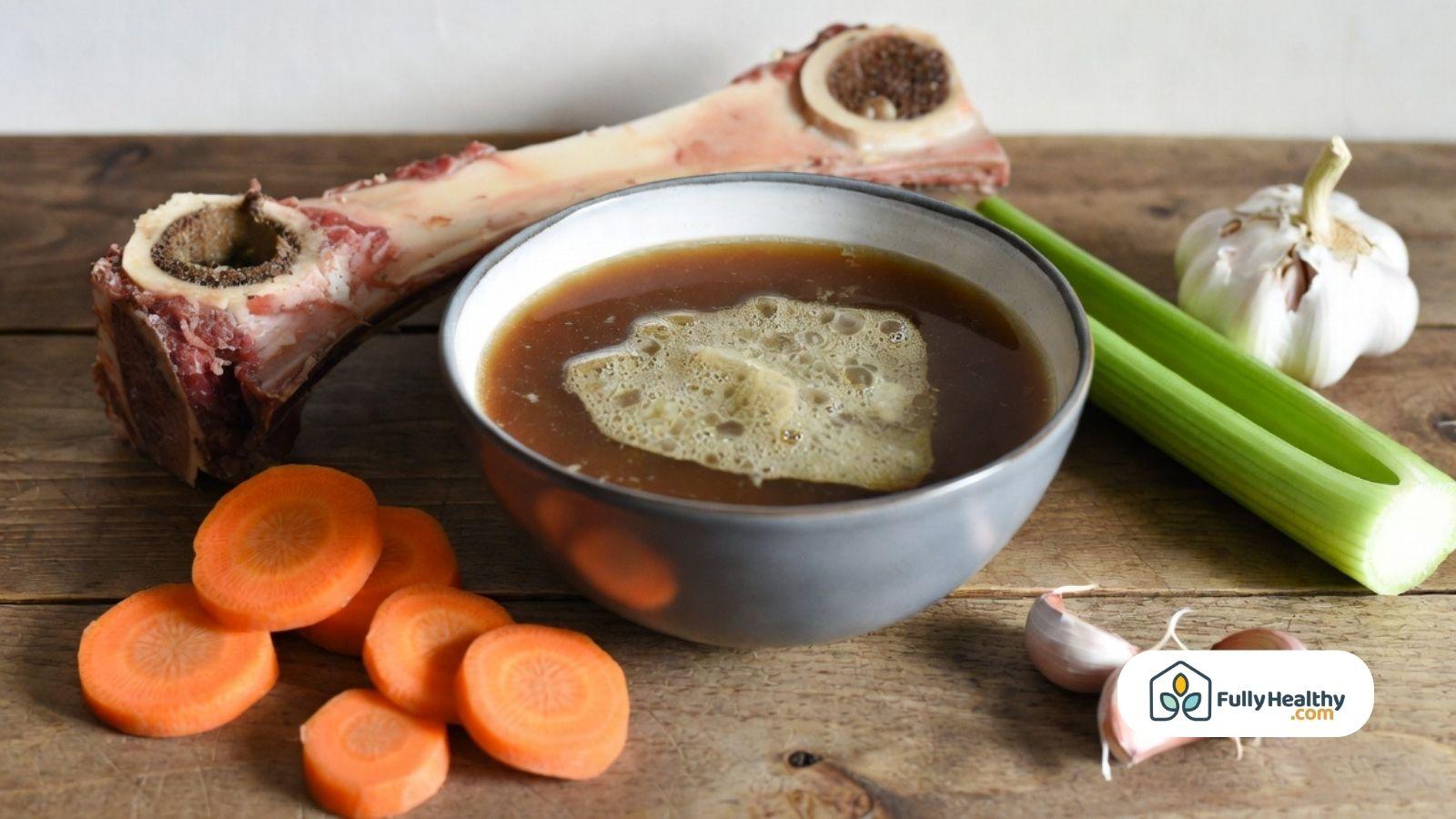 Bone broth in a bowl with beef bones, carrots, celery, and garlic on a wooden table.