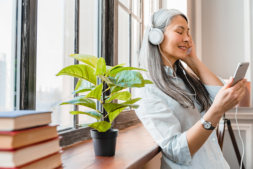 Woman with long gray hair wearing white headphones and listening to music on her cell phone. 