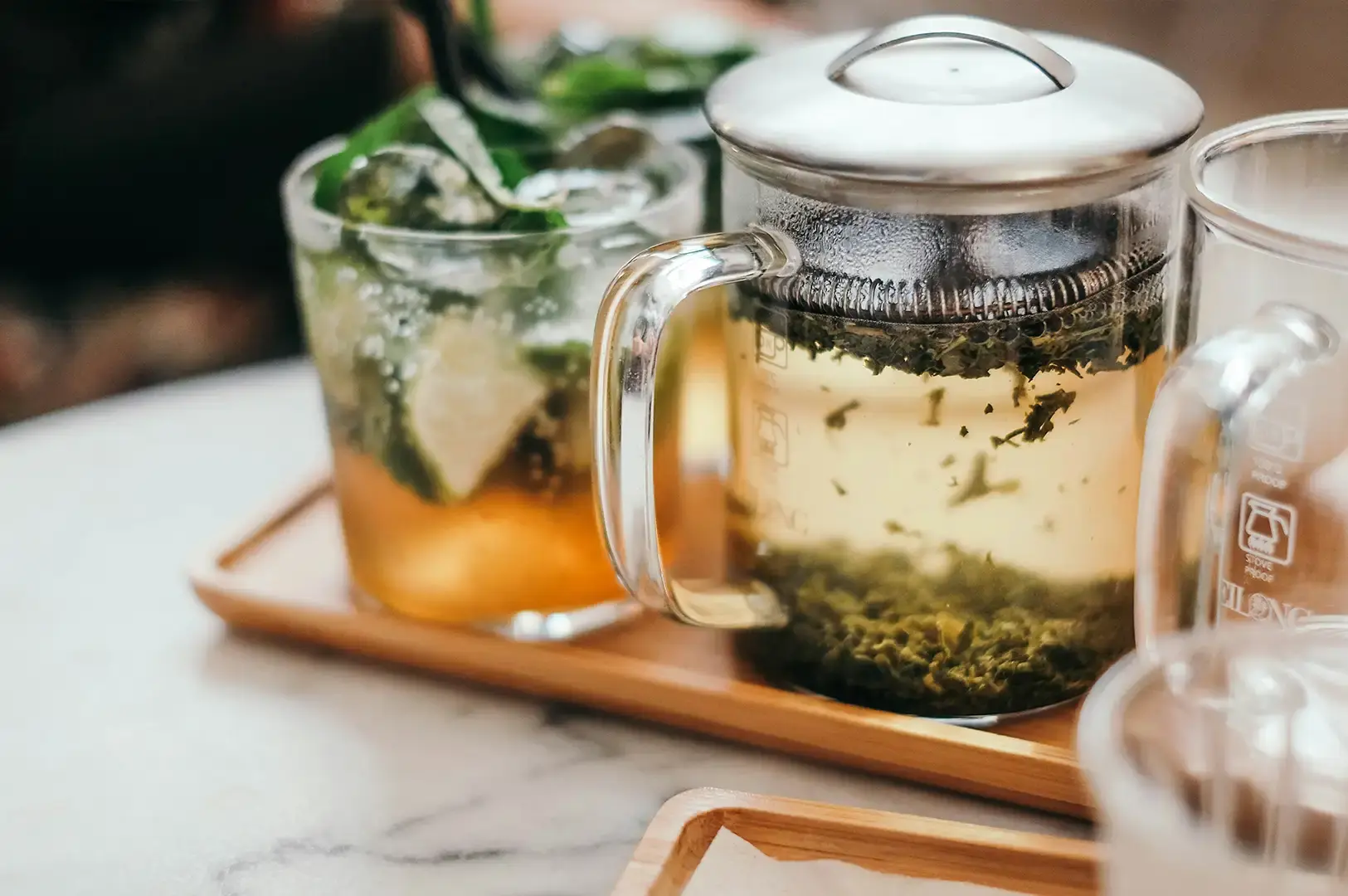 A clear glass teapot with a stainless steel filter and lid resting on a wooden tray, showing loose green tea leaves steeping and swirling in clear warm water next to an iced glass garnished with fresh green mint leaves and lime wedges.