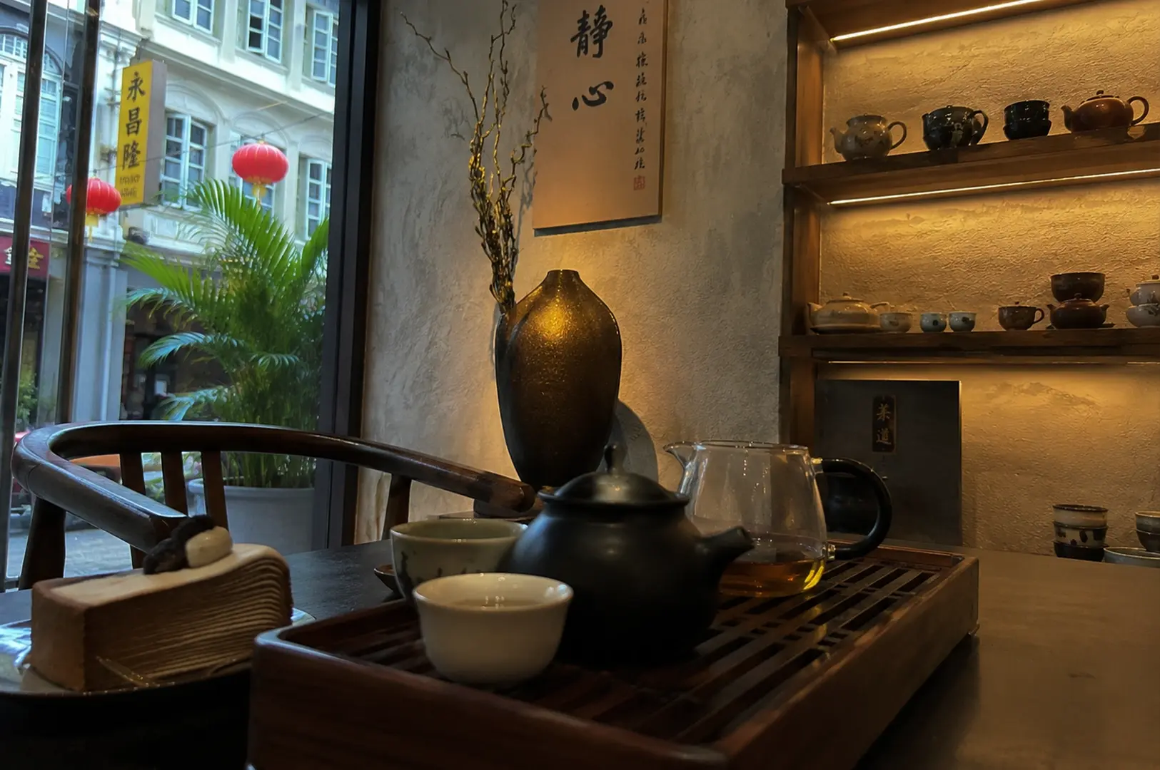 A moody, low-lit tea table setup features a dark ceramic teapot and a glass fairness cup filled with tea resting on a wooden drainage tray, with a light-colored ceramic cup in the immediate foreground and a slice of layered crepe cake served on a plate to the left. The scene is set inside a tea house with a large window displaying a vibrant city street view complete with green palms and hanging red Chinese lanterns, balanced inside by a hanging scroll with Chinese calligraphy reading "Quiet Mind" (Jing Xin) and a lit display shelf filled with various artisanal teapots.