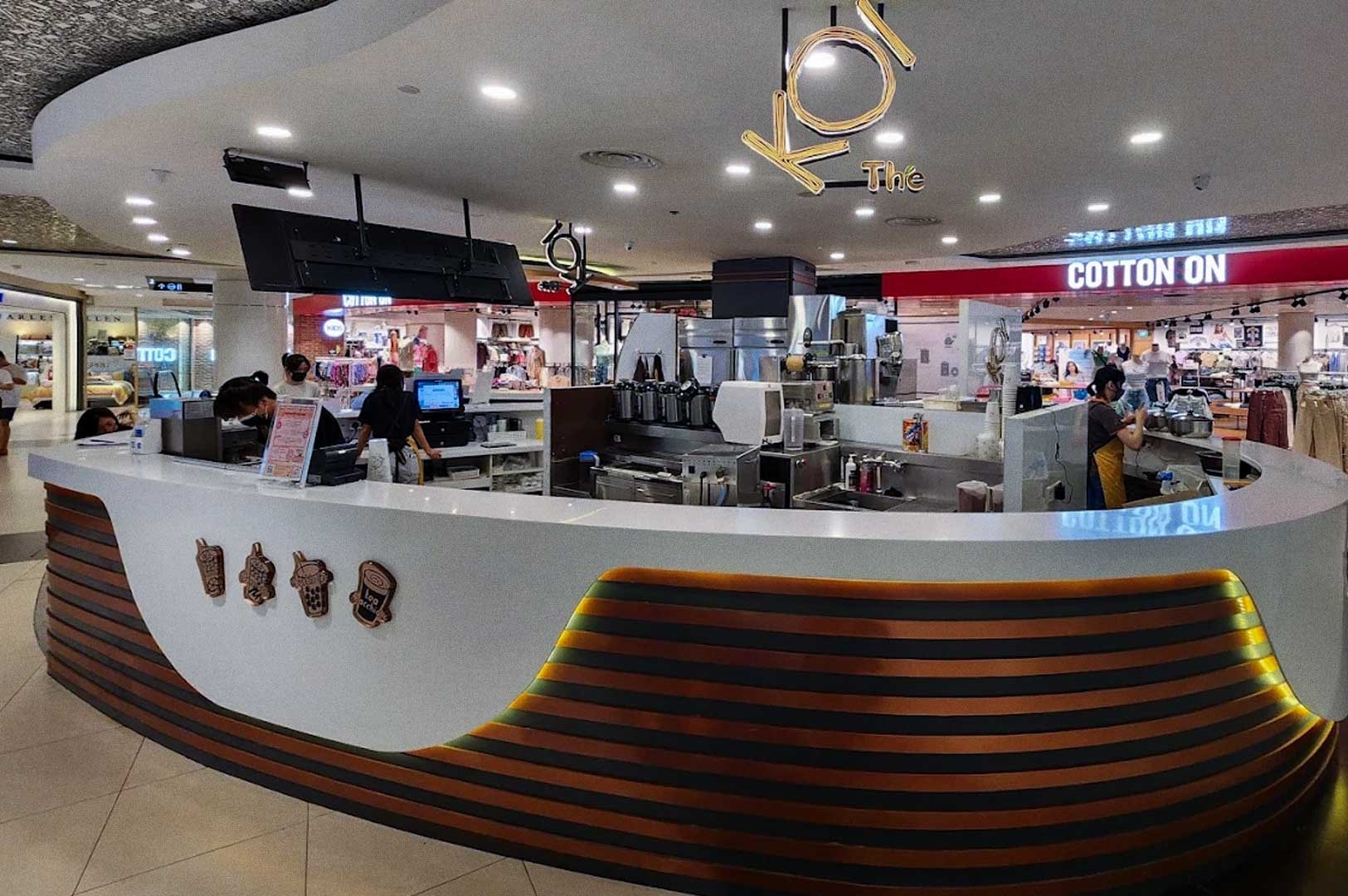 Sleek KOI Thé bubble tea counter inside a mall with a curved white and wood front, illuminated yellow lighting, staff preparing drinks, and decorative drink icons on the counter.