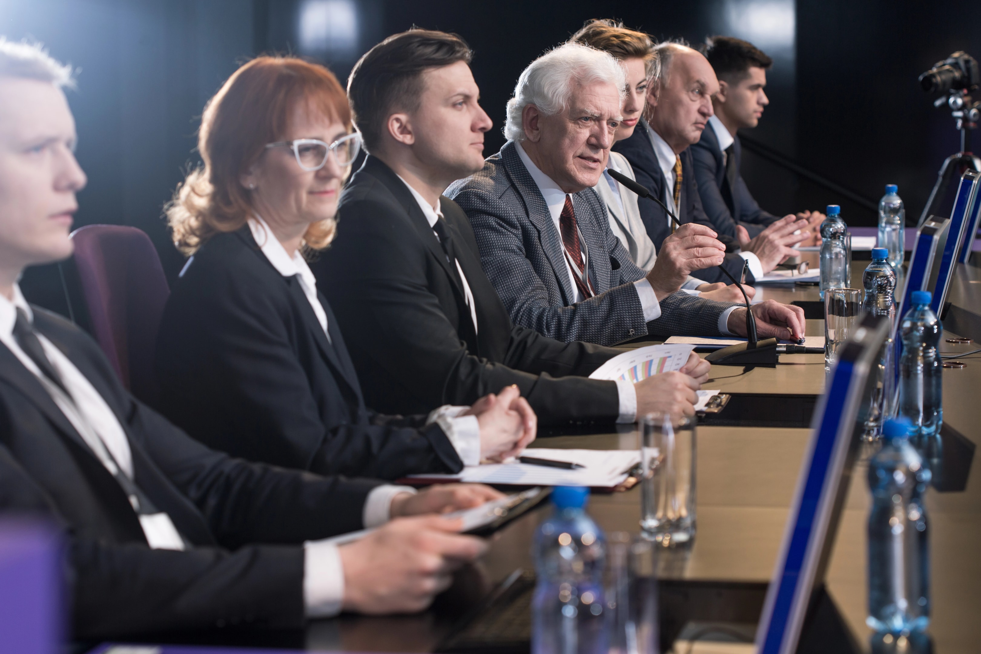 Grupa elegancko ubranych osób siedzi przy dobrze oświetlonym stole, na którym leżą notatki i stoją butelki z wodą. Jeden z mężczyzn mówi do mikrofonu, a obok stoi aparat.