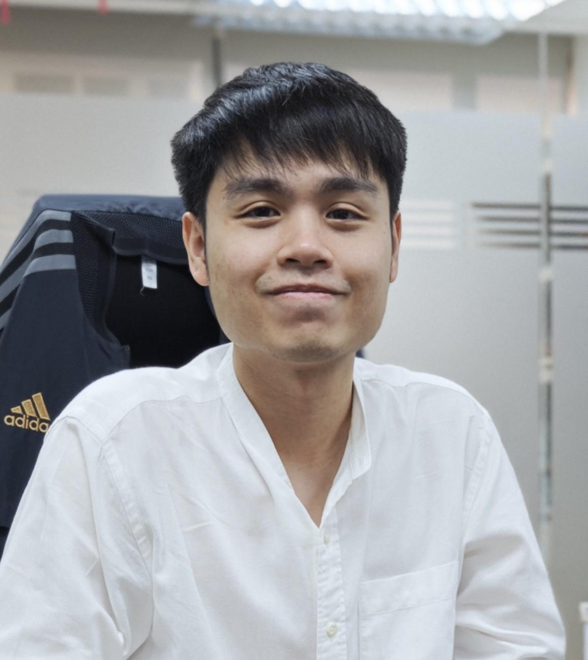 A young man in a white collarless shirt smiling warmly at the camera, seated in a casual indoor setting — conveying quiet confidence and an approachable, grounded demeanor.